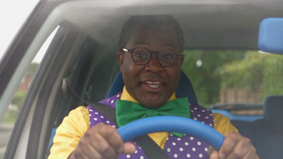 An adult with glasses and a green bow tie seated behind a blue steering wheel, wearing a yellow shirt, purple dotted vest and seat belt.